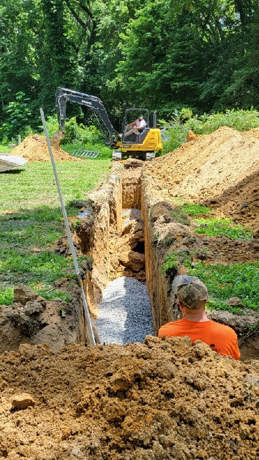 Excavator digging trench in yard with worker sitting nearby